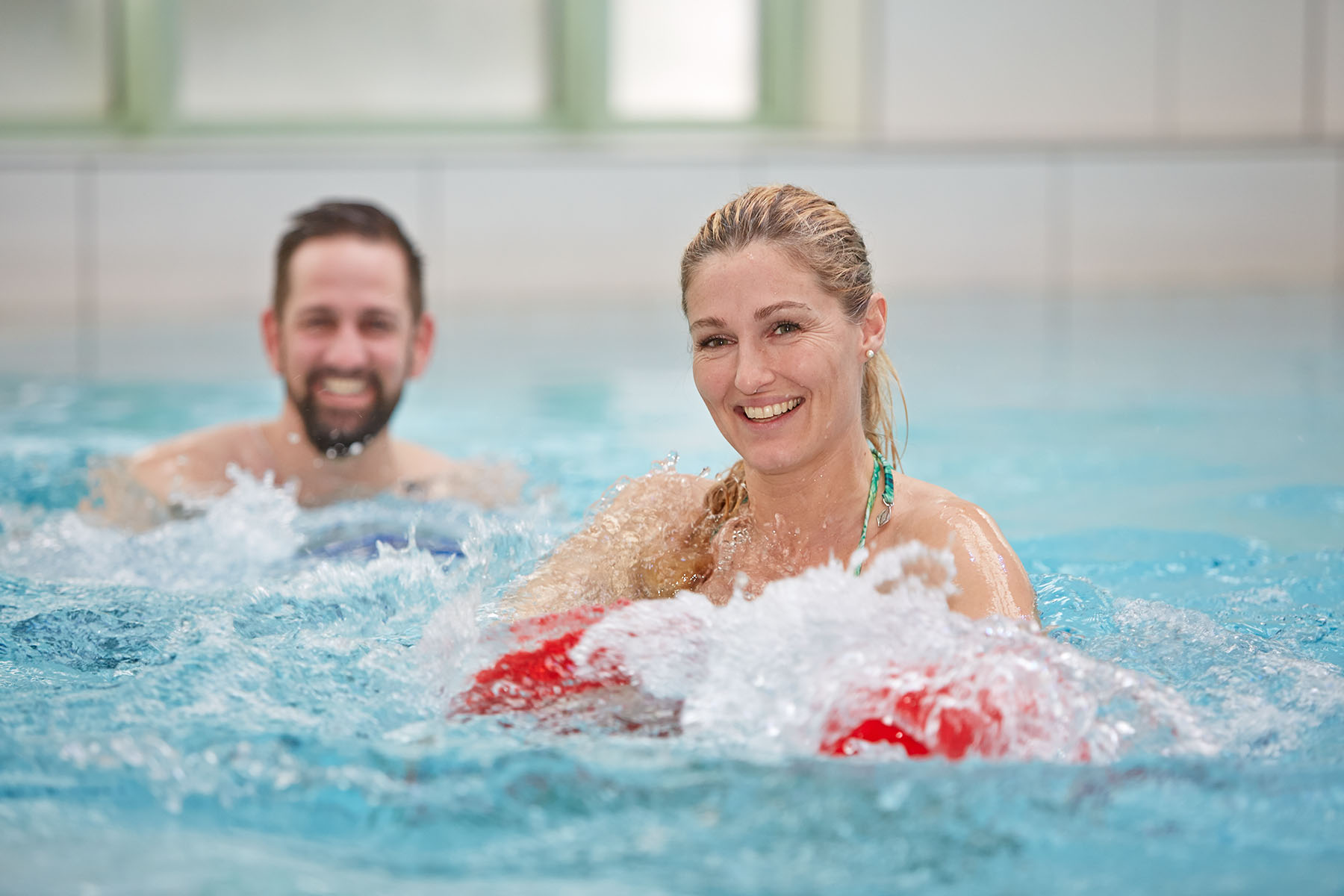 Eine Frau und ein Mann stehen in brusttiefem Wasser. Die Frau ist im Vordergrund und lächelt in die Kamera. Sie hält rote Wasserhanteln vor sich, wodurch Wasser spritzt. Der Mann befindet sich hinter ihr und ist unscharf. Beide wirken gut gelaunt. 