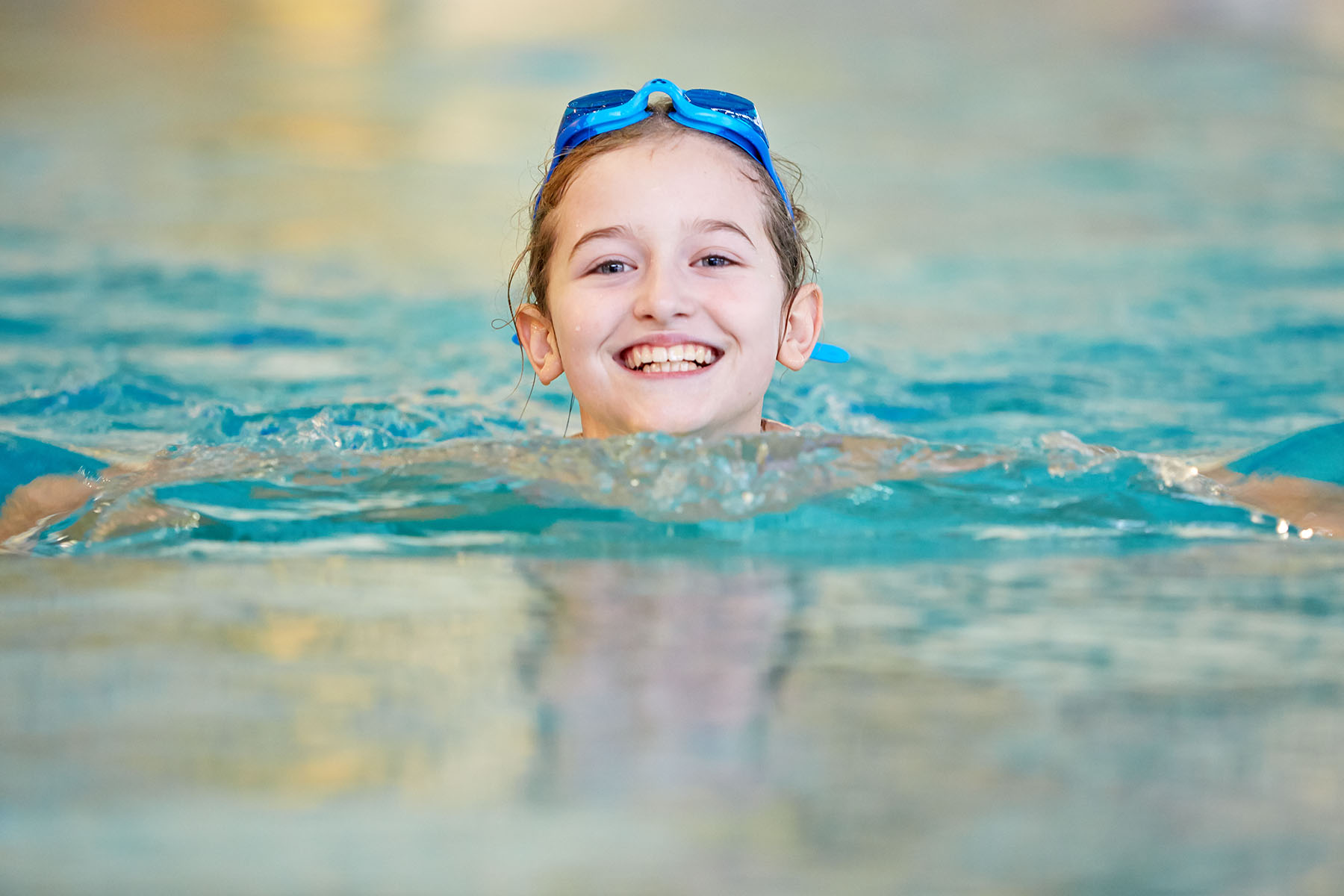 Ein Kind schwimmt im Hallenbad und lächelt in die Kamera. Der Kopf ist über Wasser, auf dem Kopf liegt eine blaue Schwimmbrille. Das Wasser ist hellblau, der Hintergrund unscharf. Die Stimmung wirkt freundlich und fröhlich. 