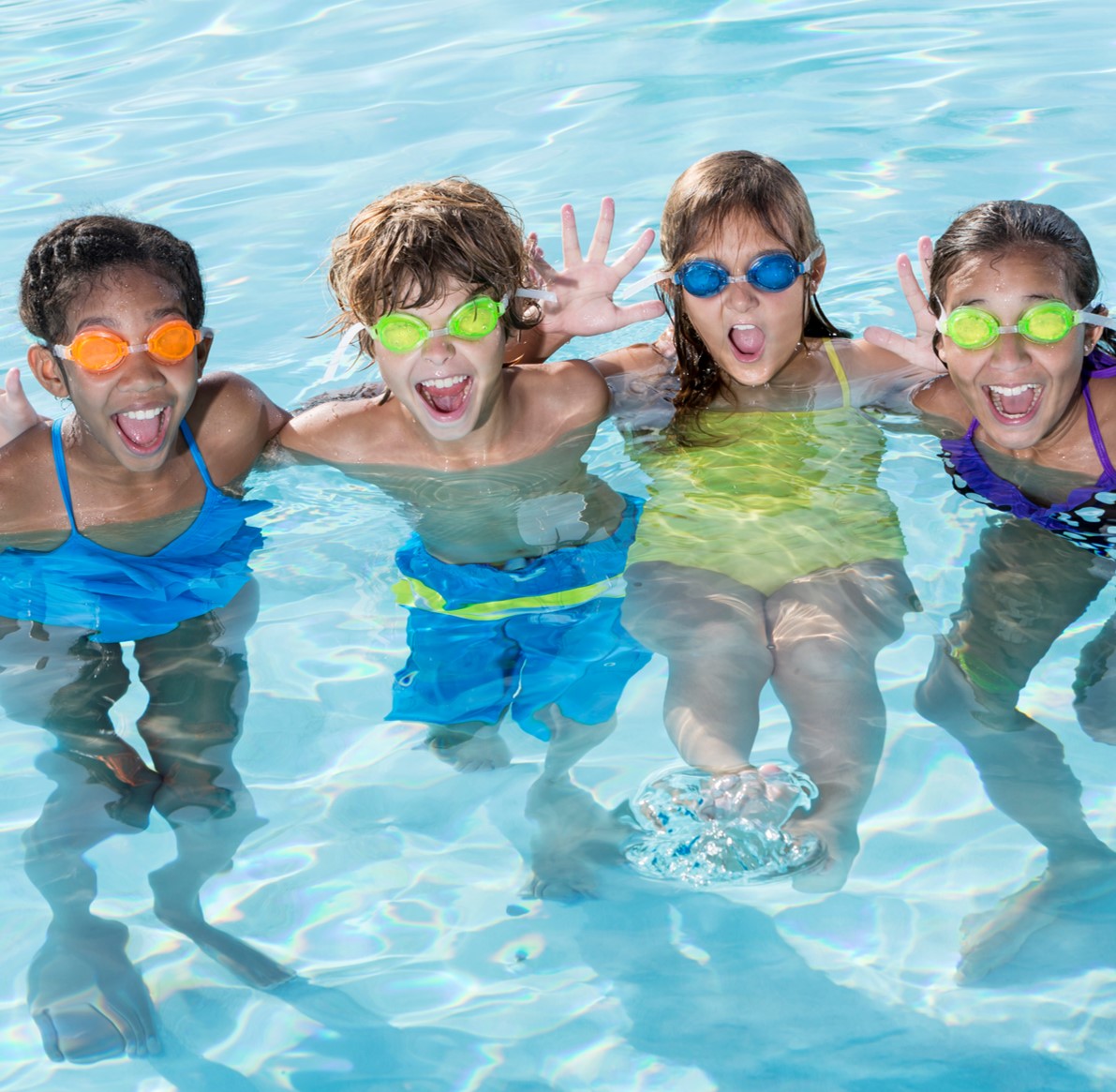 Vier Kinder stehen lachend im Wasser eines Schwimmbeckens. Sie tragen bunte Schwimmbrillen und Badekleidung, schauen zur Kamera und wirken fröhlich. Das Wasser ist klar und hellblau, die Stimmung spielerisch und lebendig.