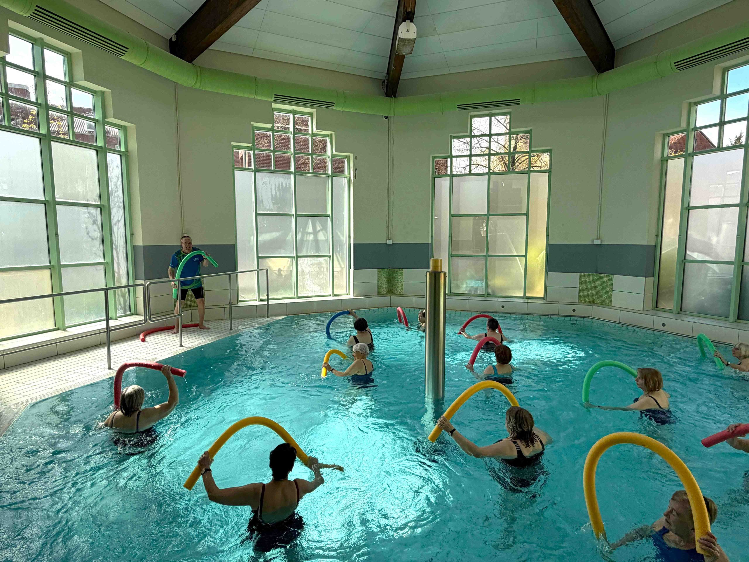 Mehrere Senioren stehen in einem Hallenbad im Wasser und machen Übungen mit bunten Schwimmnudeln. Eine Person am Beckenrand leitet die Gruppe.
